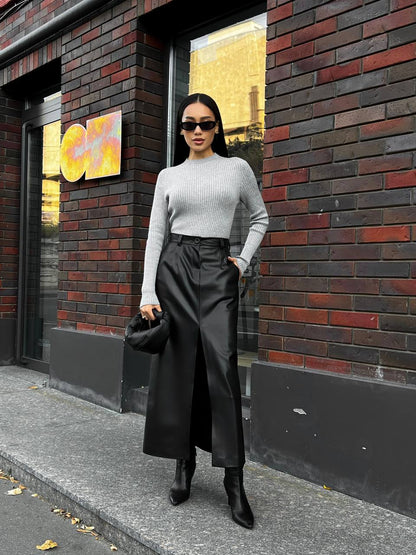 Woman in a light gray sweater and black pants standing against a brick wall.