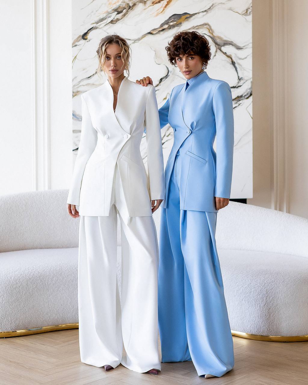 Two women in formal suits standing in a stylish interior setting.