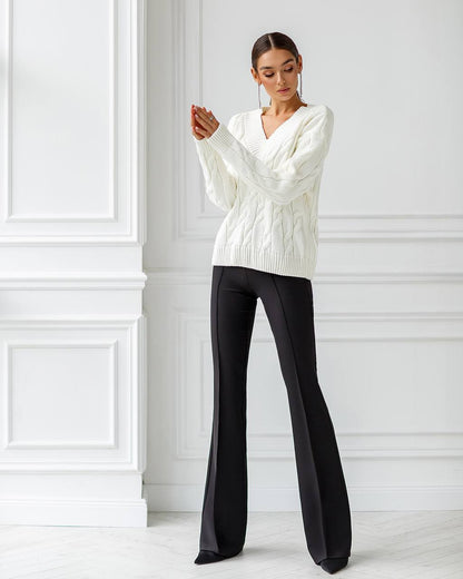 Woman wearing a white cable knit sweater and black pants against a white wall.