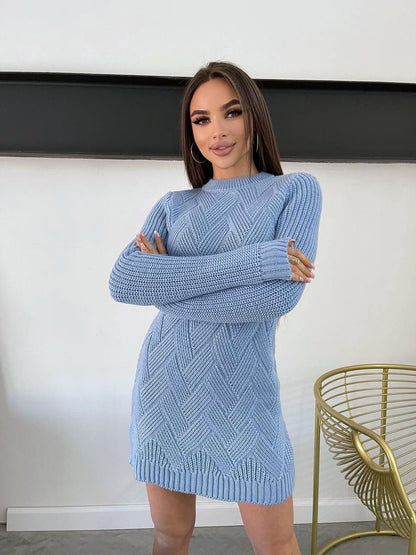 Woman wearing a blue knitted dress standing indoors with a white wall and gold chair in the background.