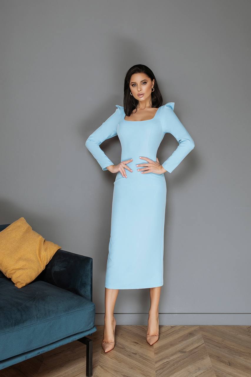 Woman in a light blue dress standing in a room with a gray wall and a blue couch.