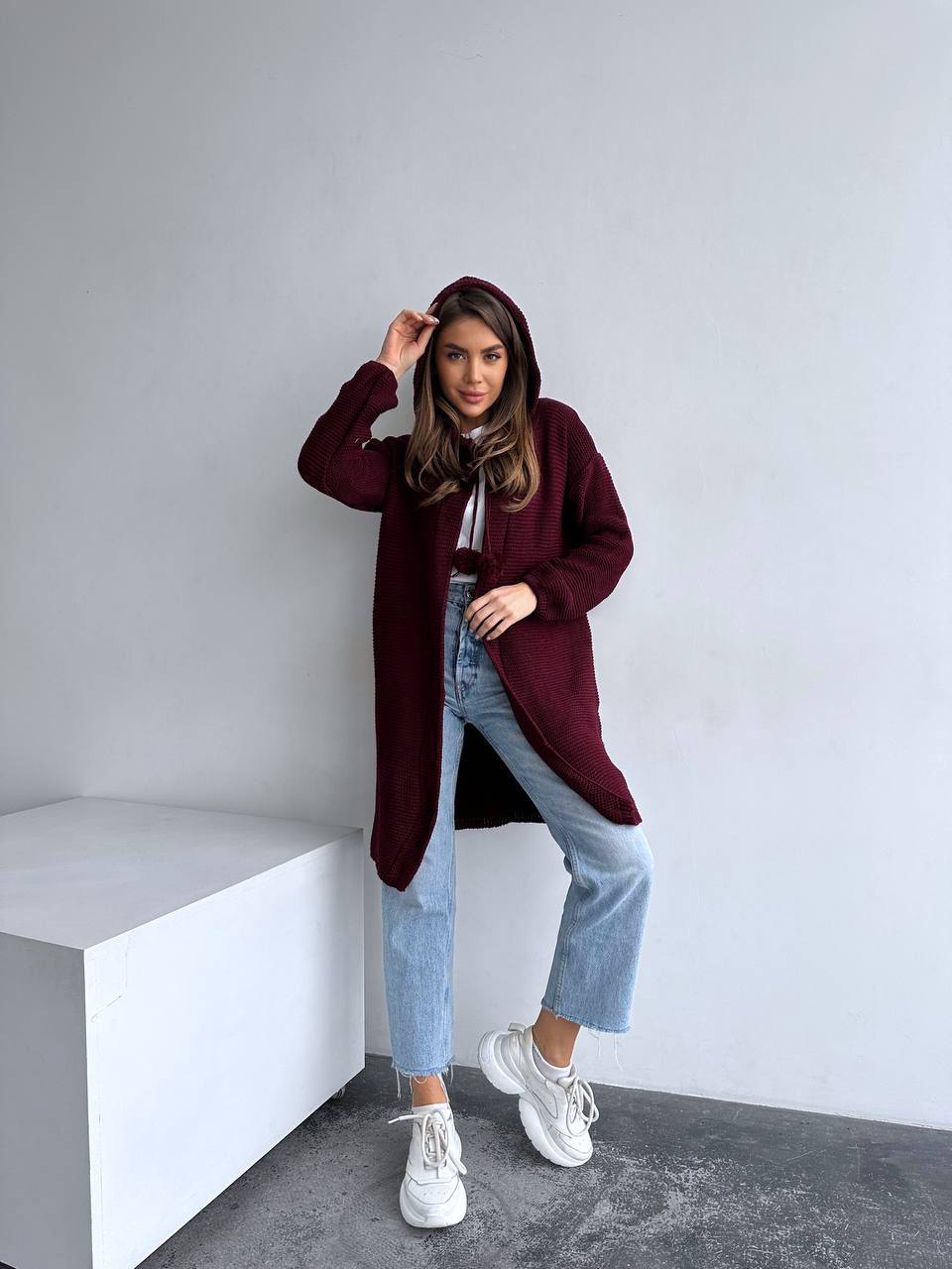 Woman wearing a burgundy coat, white shirt, blue jeans, and white sneakers in a minimalistic setting.