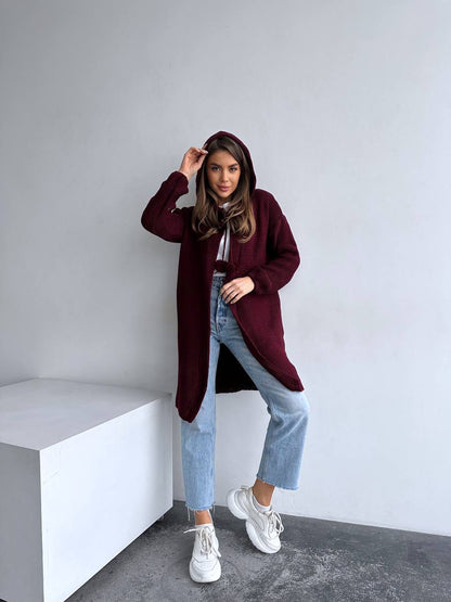 Woman wearing a burgundy coat, white shirt, blue jeans, and white sneakers in a minimalistic setting.