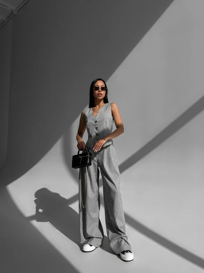 Woman in a light gray sleeveless jumpsuit standing against a gray wall with shadows.