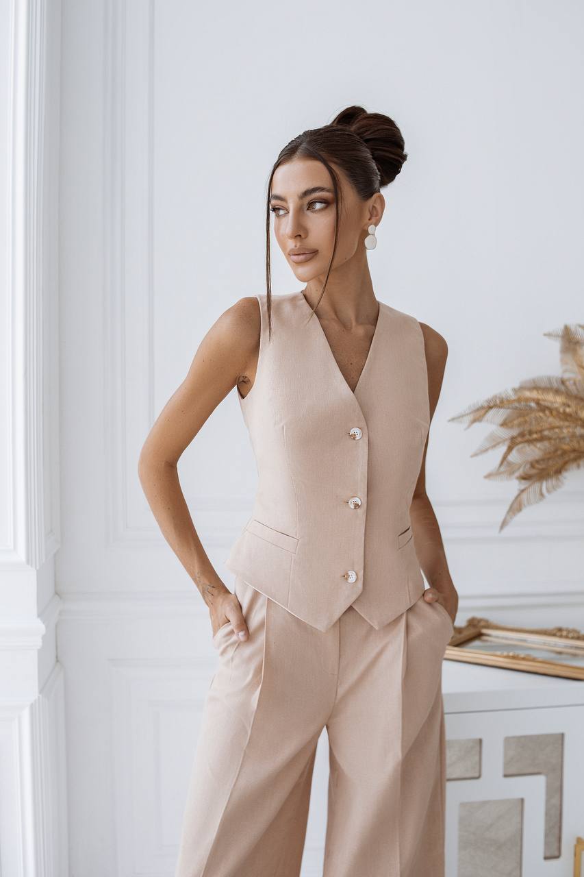 Woman wearing a beige vest and pants set in an indoor setting with decorative elements.