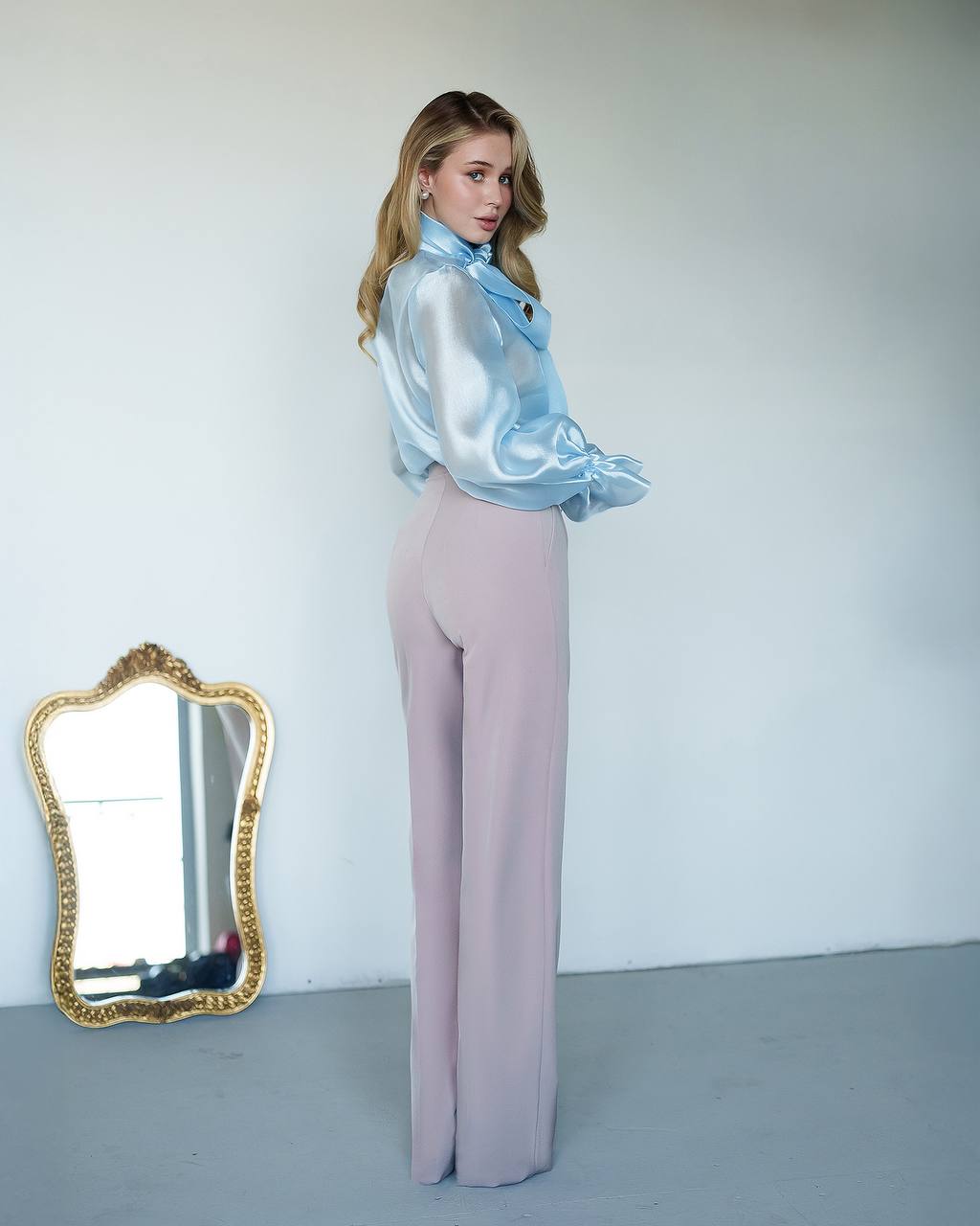 Woman in a light blue satin blouse and pink pants standing next to a decorative mirror.