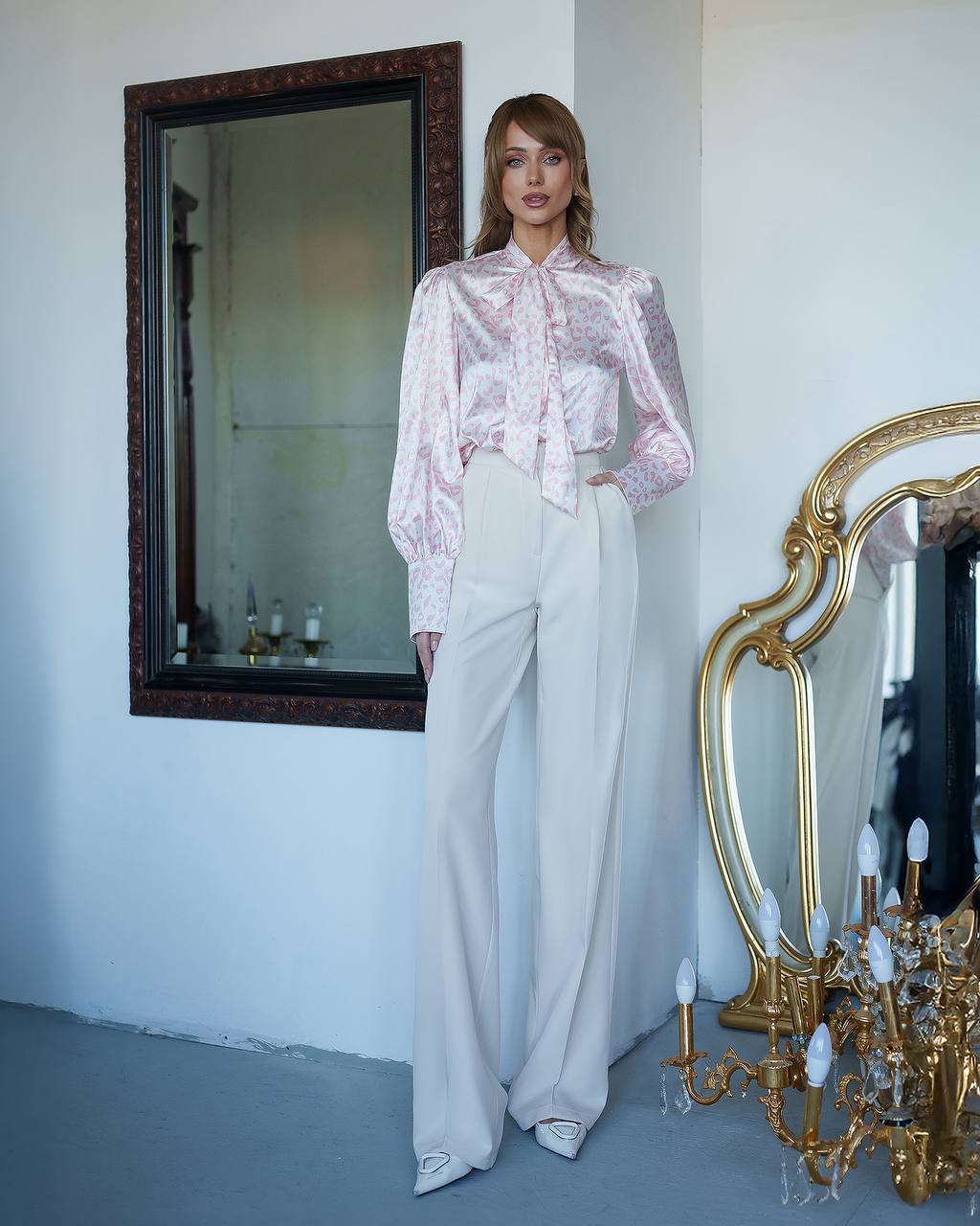 Woman in a pink blouse and white pants standing in a room with a mirror and chandelier.