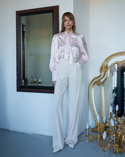 Woman in a pink blouse and white pants standing in a room with a mirror and chandelier.