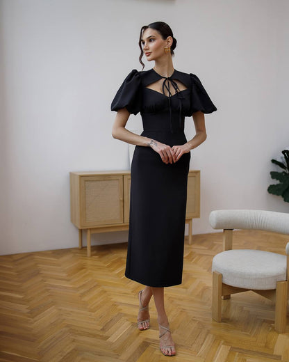 Woman in a black dress standing in a room with wooden flooring and a white wall.