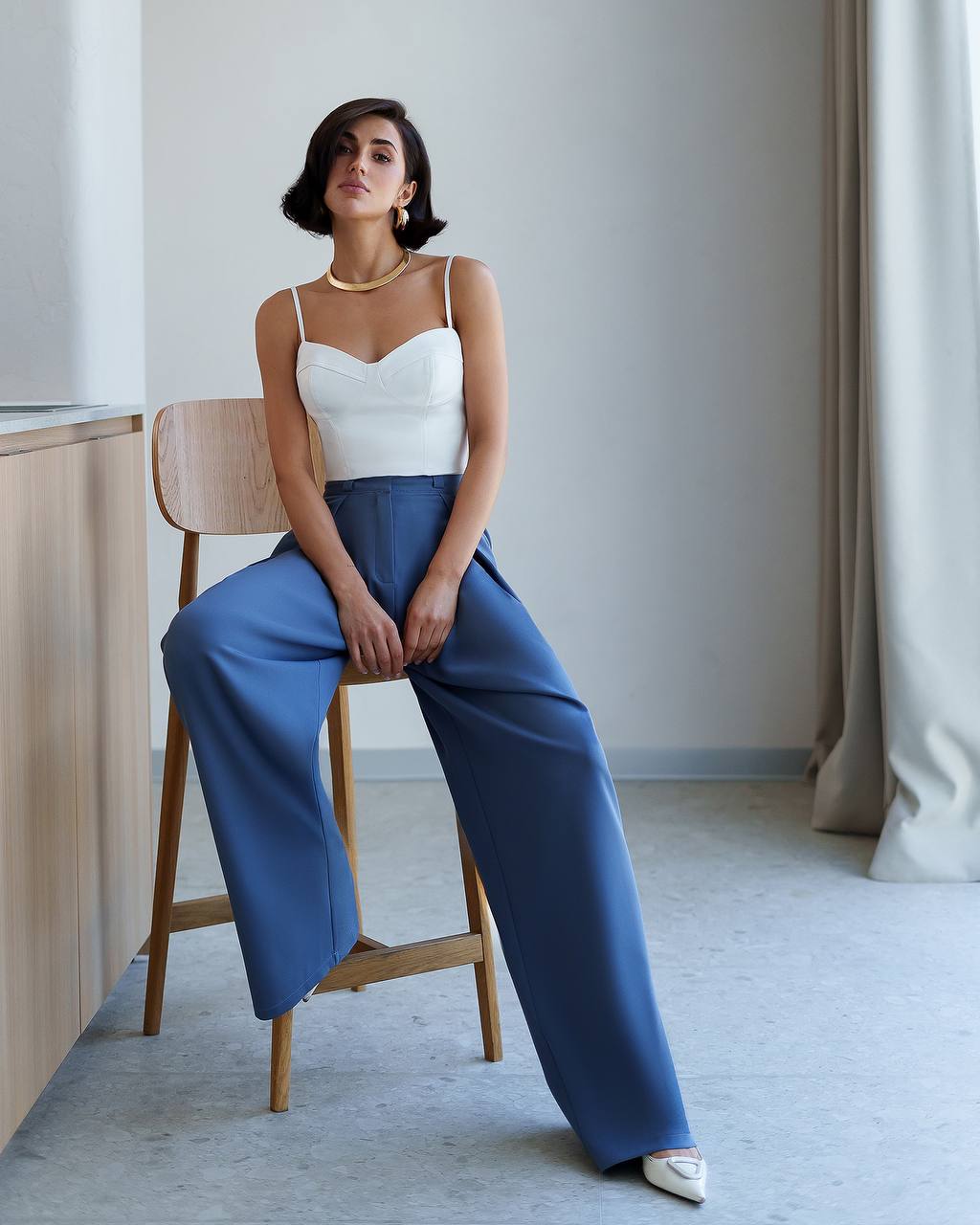 Woman sitting on a wooden chair wearing a white top and blue pants in a minimalistic room.
