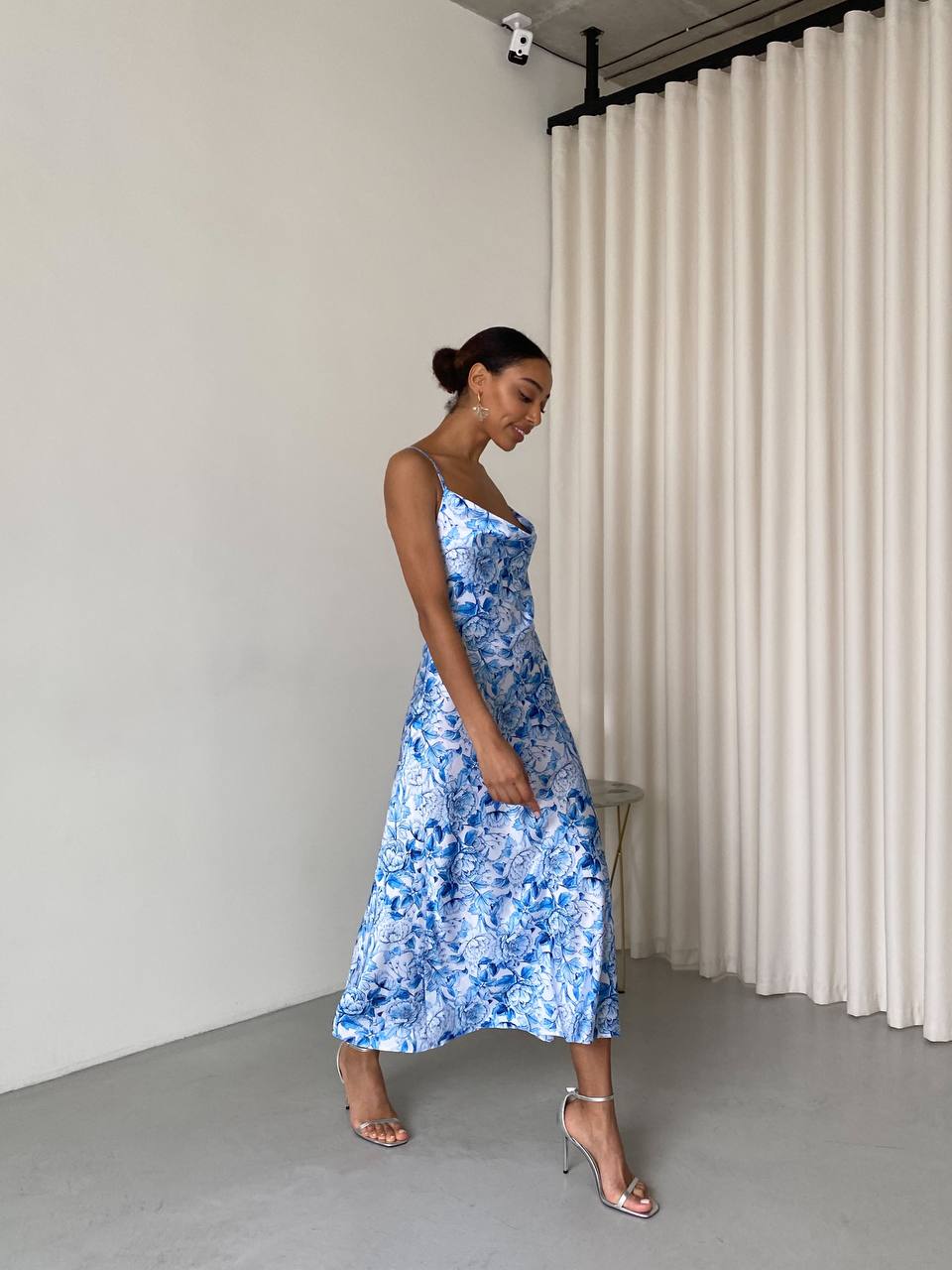 Woman wearing a blue floral dress standing in a minimalistic room with white curtains.