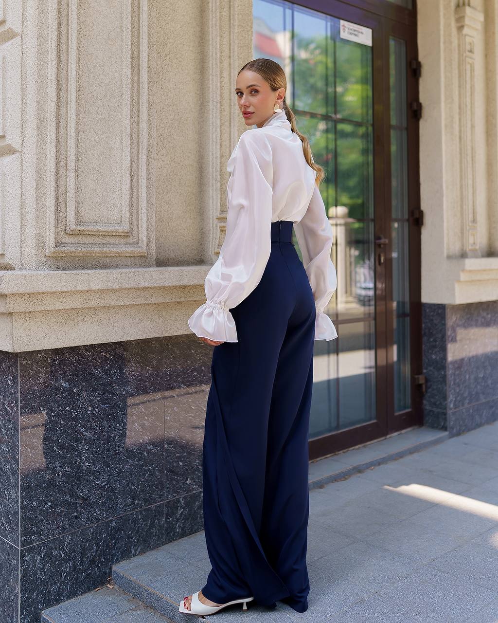 Woman in a white blouse and navy pants standing in front of a building entrance.