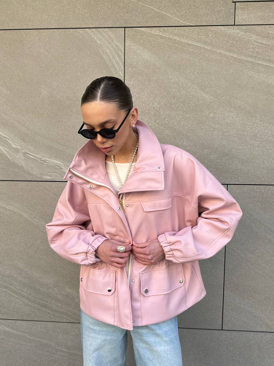 Person wearing a pink jacket and sunglasses against a gray wall