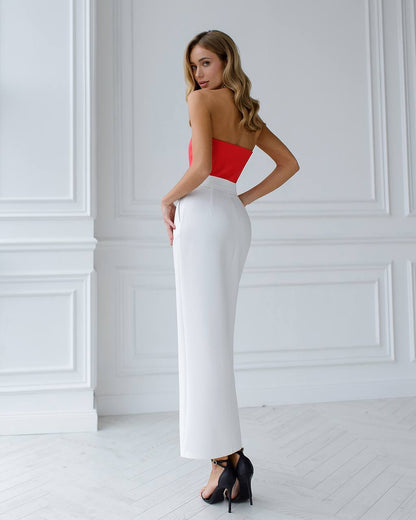 Woman wearing a red top and white pants against a white wall.
