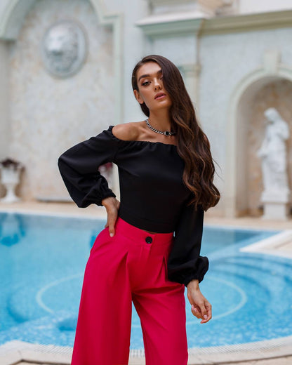 Woman in black off-shoulder top and red pants standing by a pool with classical architecture in the background