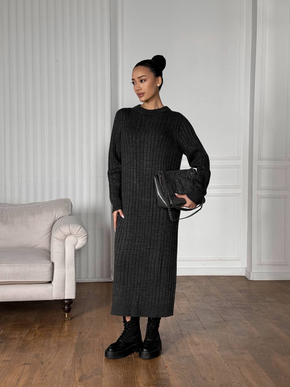Woman in a black ribbed dress holding a black clutch in a room with a gray sofa and white paneled walls.