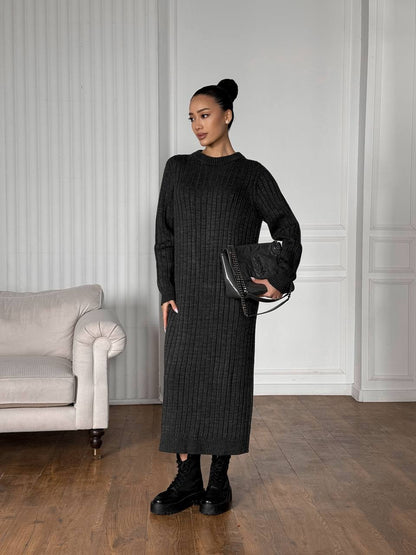 Woman in a black ribbed dress holding a black clutch in a room with a gray sofa and white paneled walls.