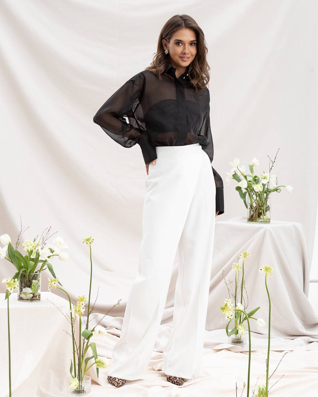 Woman in black top and white pants standing in a minimalistic setting with flowers.
