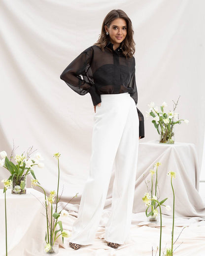 Woman in black top and white pants standing in a minimalistic setting with flowers.