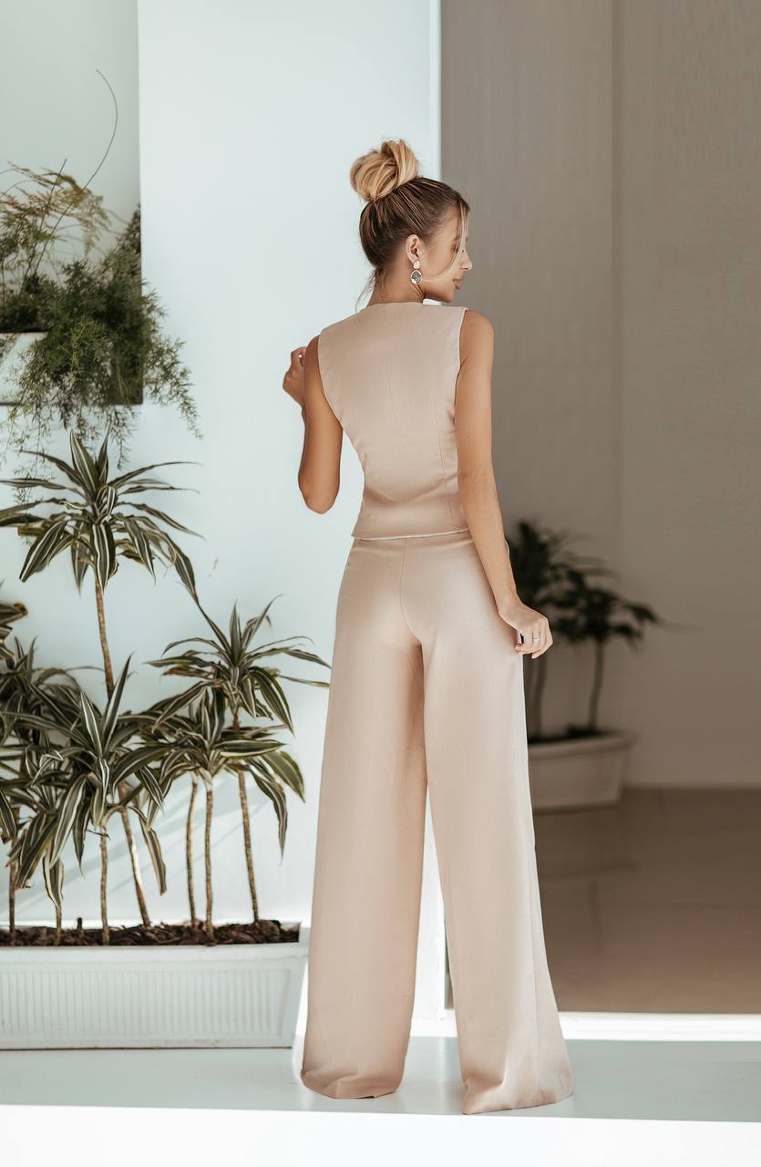 Woman in a beige jumpsuit standing in a room with plants