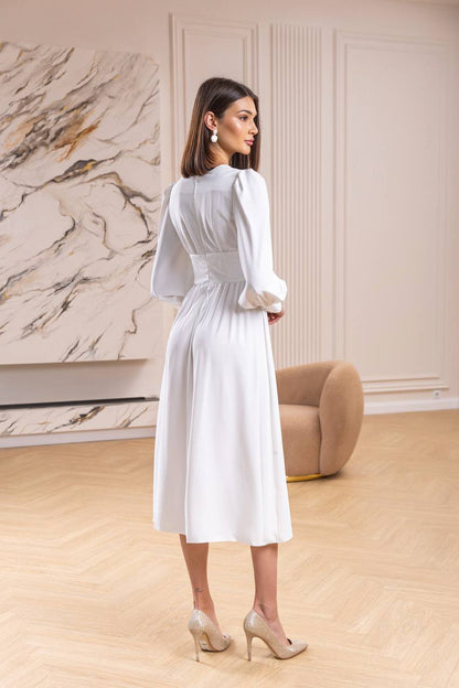 Woman in a white dress standing in a stylish room with marble wall and beige armchair.