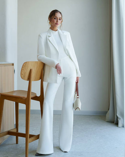 Woman in a white suit standing in a minimalistic room with a wooden chair and light-colored walls.