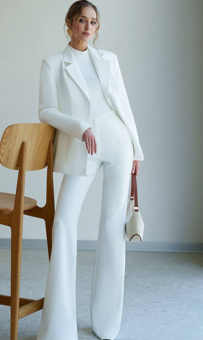 Woman in a white suit standing next to a wooden chair against a plain background