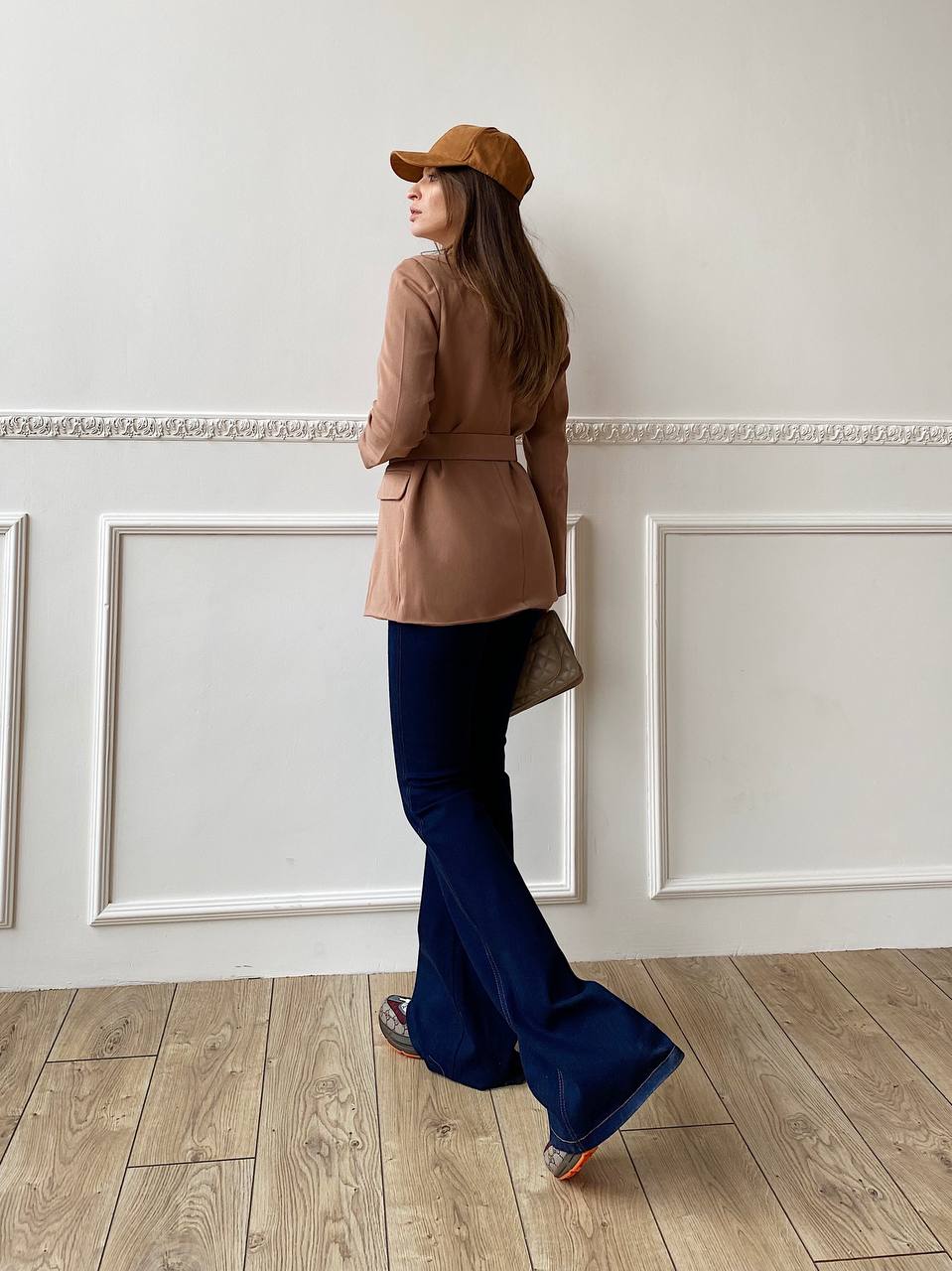 Woman in a beige jacket and blue flared pants standing in a room with white walls and wooden floor.