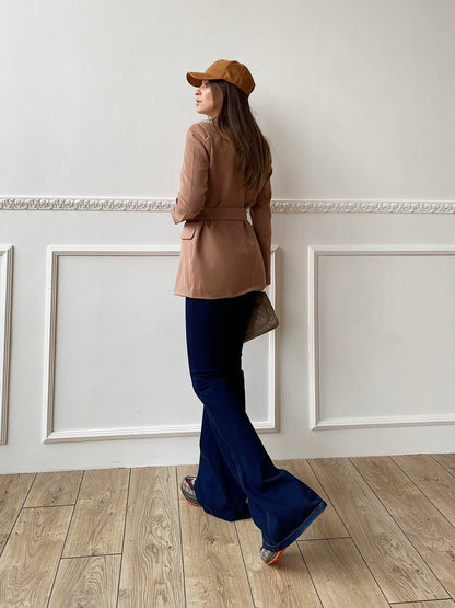 Woman in a beige jacket and blue flared pants standing in a room with white walls and wooden floor.