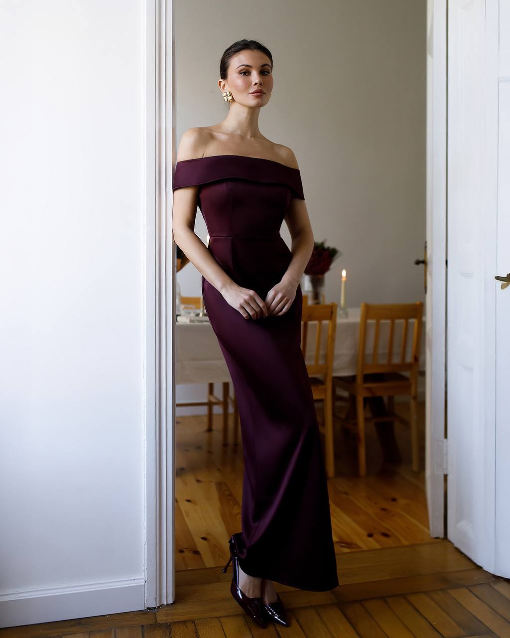 Woman in a dark purple off-shoulder gown standing in a room with wooden flooring and a dining table.