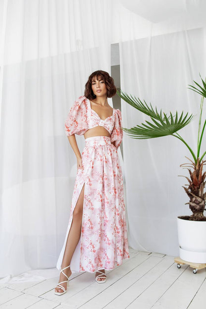 Woman in a floral dress standing in a bright room with white curtains and a plant.