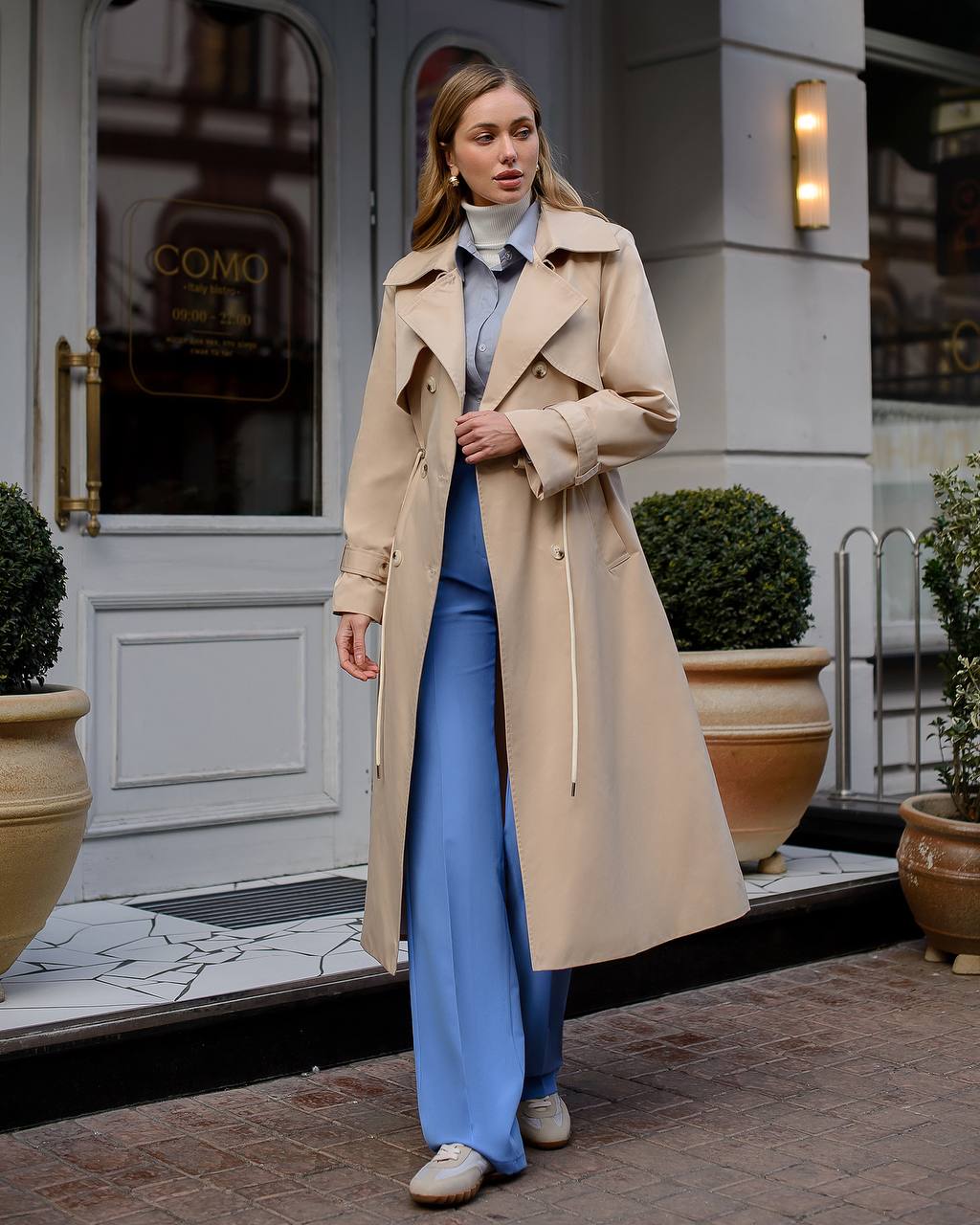 Woman in a beige trench coat and blue outfit standing in front of a building with 'COMO' on the door.