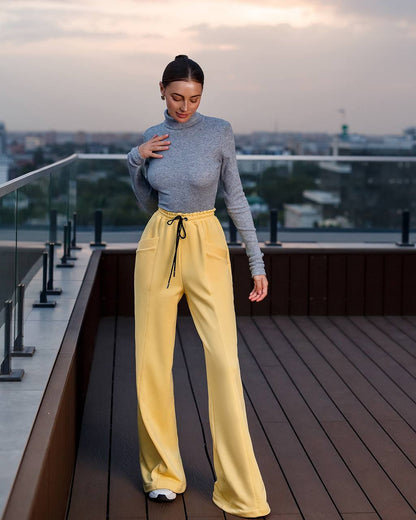 Woman wearing a gray sweater and yellow pants on a rooftop with a cityscape background