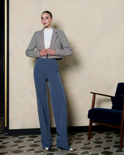 Woman in a checkered blazer and blue pants standing in a room with a chair and patterned floor.