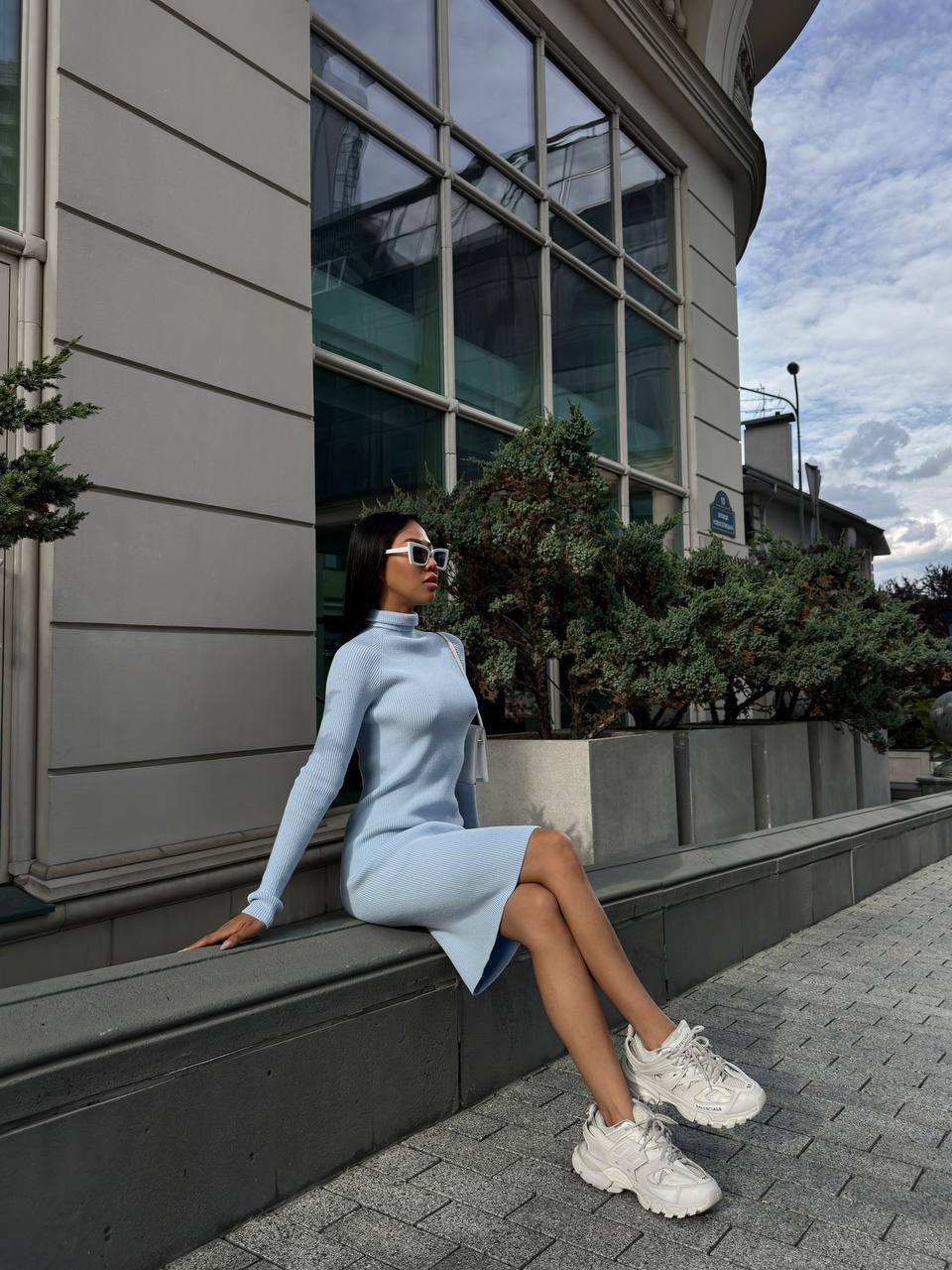 Woman in a light blue dress and white sneakers sitting on a building ledge with a cityscape background.