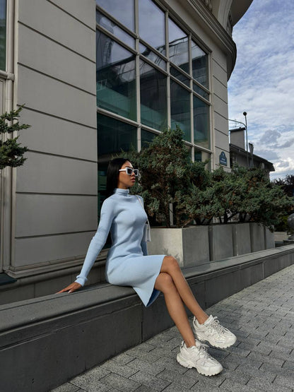 Woman in a light blue dress and white sneakers sitting on a building ledge with a cityscape background.