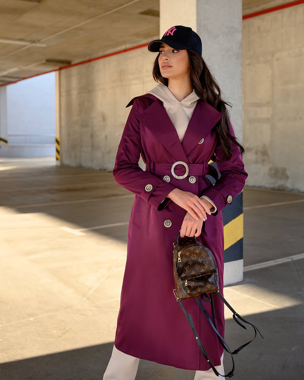 Woman wearing a purple coat and black cap holding a brown handbag in an outdoor setting.