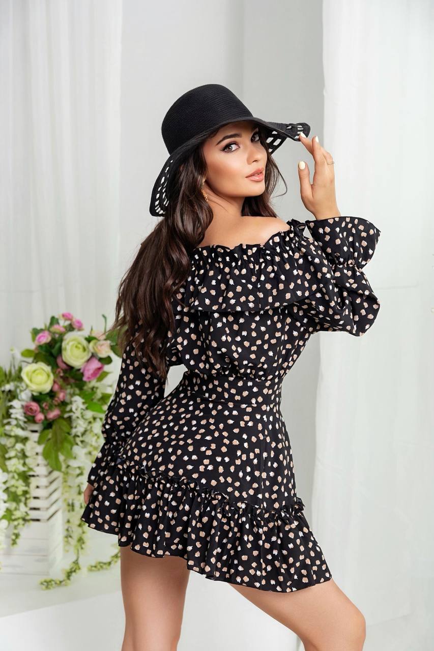 Woman wearing a black polka dot dress and black hat against a white curtain background