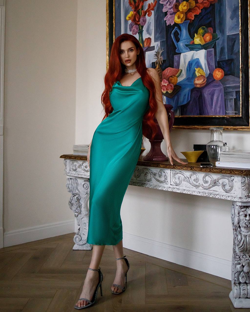 Woman in a teal dress standing in a room with a decorative table and colorful painting.