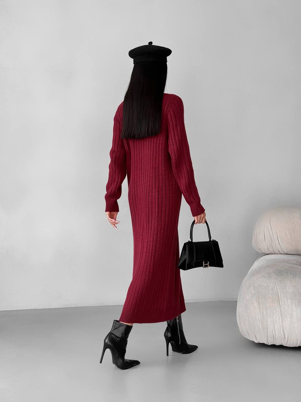 Person wearing a burgundy ribbed dress and black beret, holding a black handbag against a plain background.