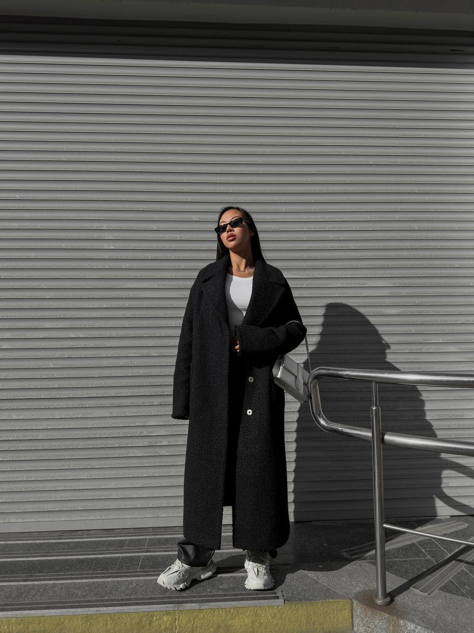 Person wearing a long black coat standing in front of a closed metal shutter.
