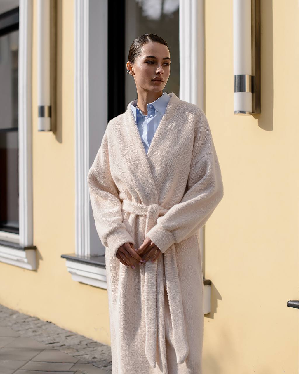 Woman wearing a beige robe standing outside a building with yellow walls.