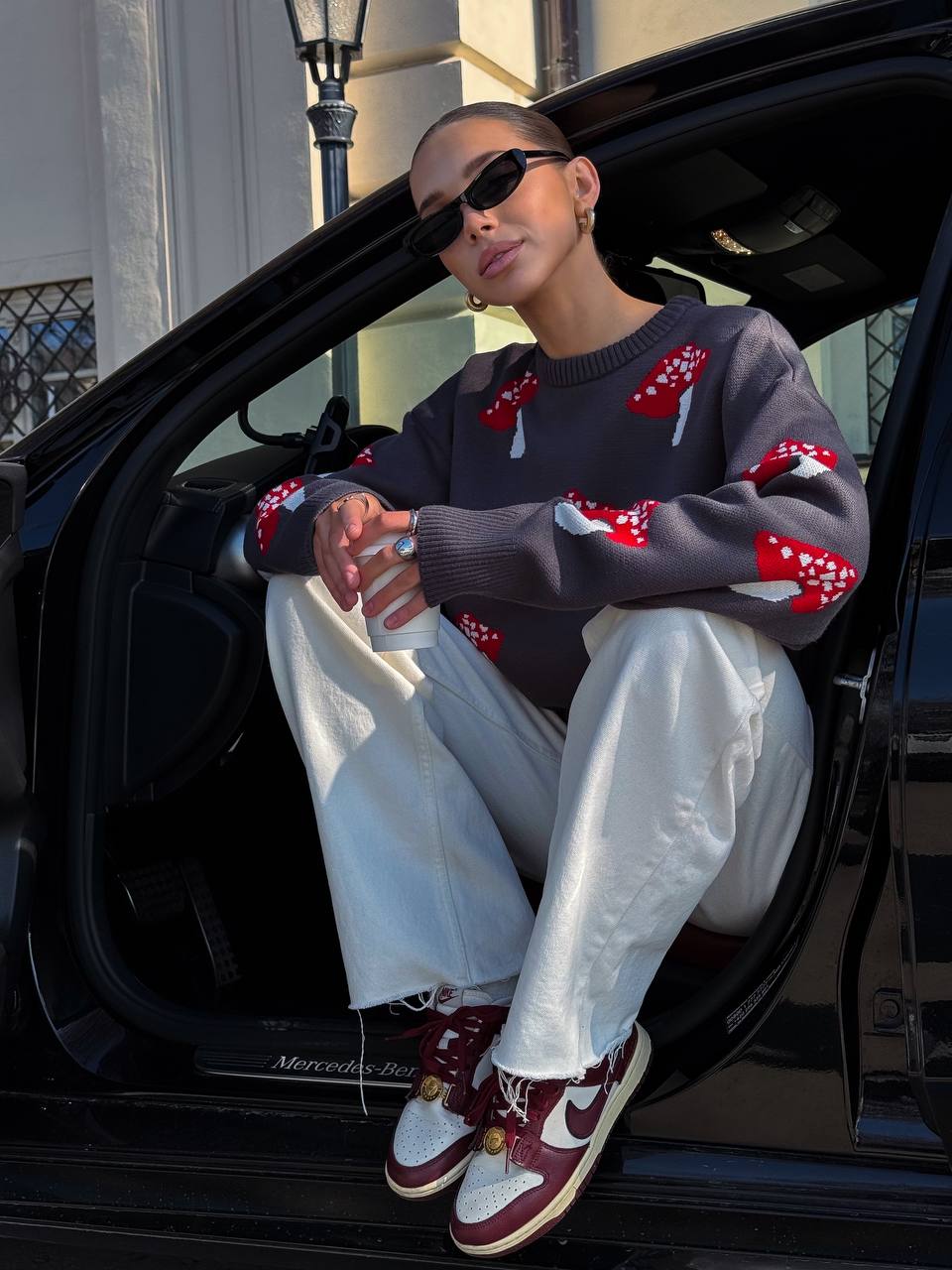 Person sitting in an open car door wearing a patterned sweater and white pants.