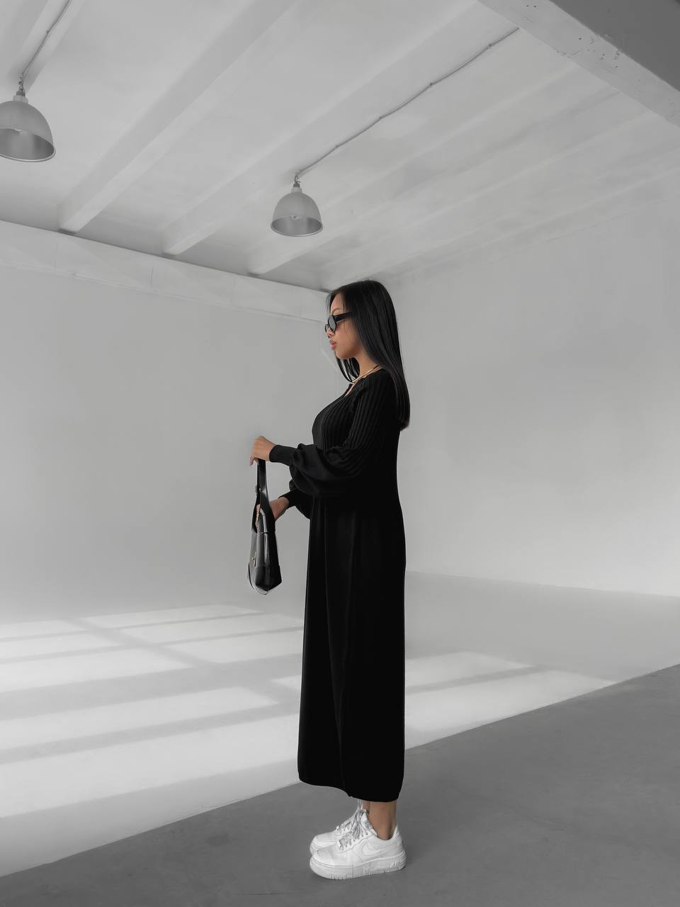 Woman in a black dress holding a black bag in a minimalistic white room.