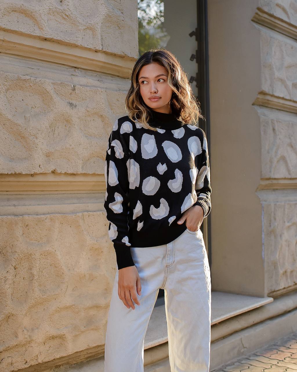 Woman wearing a black sweater with white patterns and white pants standing in front of a stone building.