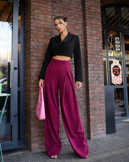 Woman in black top and magenta pants holding a pink bag against a brick wall.