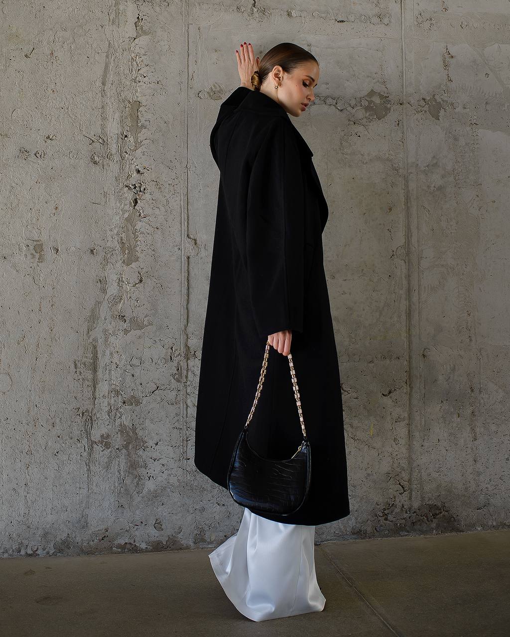 Person wearing a black coat and white pants holding a black handbag against a textured wall.