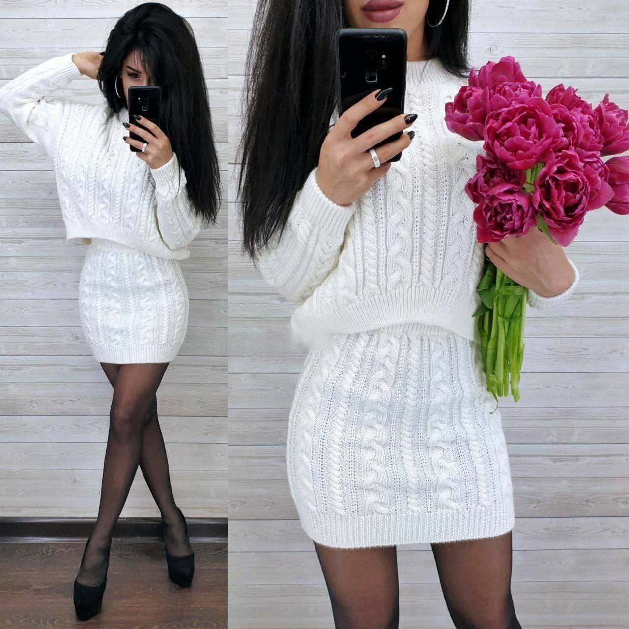 Woman in a white cable knit dress holding pink flowers, taking a mirror selfie.