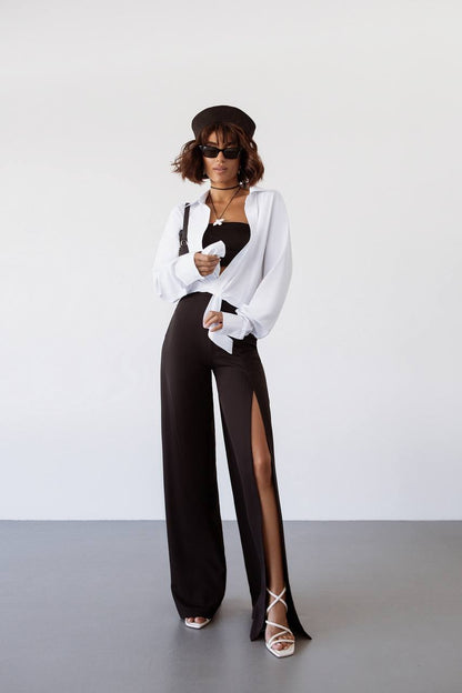 Woman wearing a white blouse, black pants with a high slit, and sunglasses on a plain background