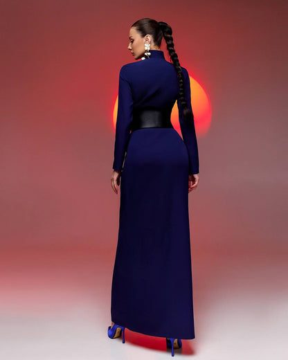 Woman in a long blue dress with a high collar and black belt, standing against a gradient red to pink background.